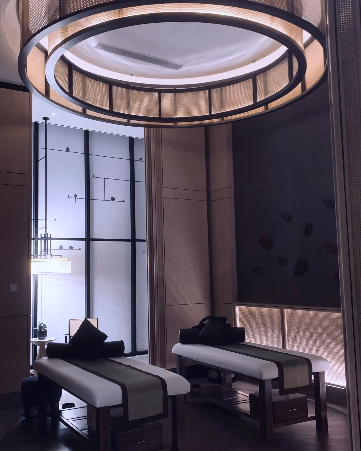 A treatment room at Marin Aesthetics: two made-up treatment beds beneath a softly lit circular pendant, framed by warm wood and woven screens